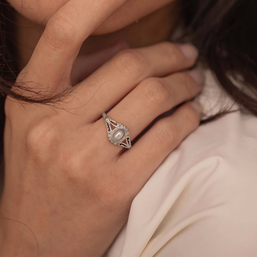 model image - A silver ring with an oval white pearl and small clear stones, worn on a person's hand against a soft cream-colored top.