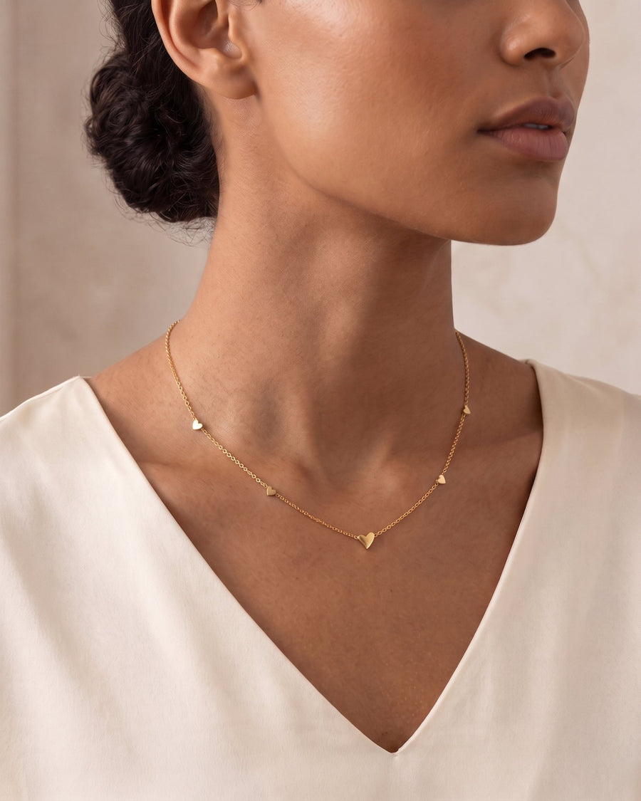 Model Image - A close-up of a model wearing a delicate gold chain necklace with small heart charms, paired with a cream-colored V-neck top.