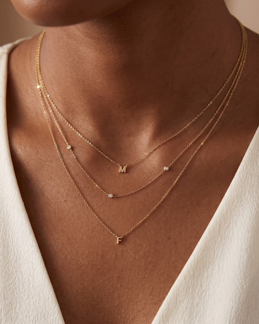 Model Image - A close-up of a model wearing a layered gold necklace set with small letter pendants and tiny clear stones, paired with a white V-neck top.