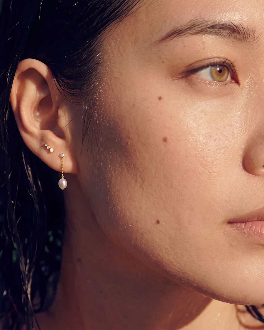Model Image - A close-up of a woman's ear wearing a delicate gold ear cuff with small clear stones and a dangling white pearl earring, shown outdoors with wet hair.