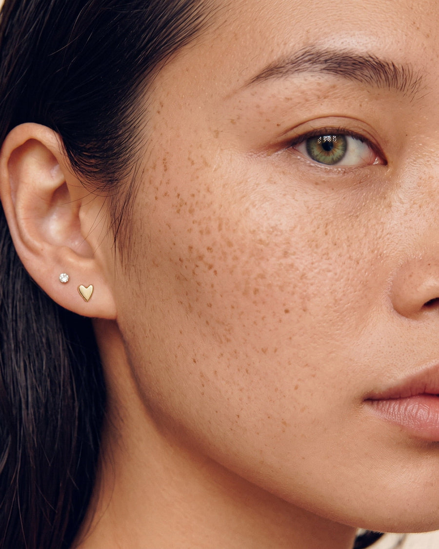 Model image - Close-up of a woman's ear wearing two small stud earrings, one gold heart-shaped and one silver oval, against a light beige background.