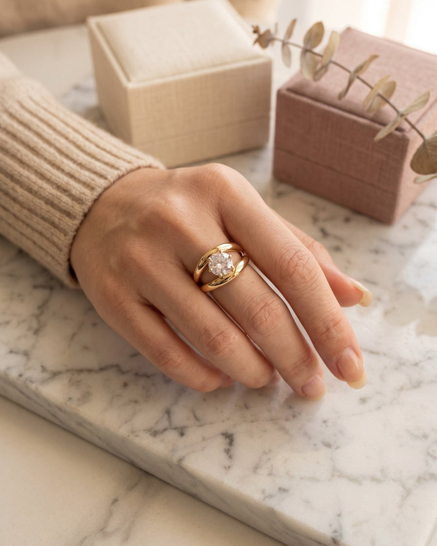 model image - A gold ring with a clear central gemstone worn on a person's hand, resting on a white marble surface with jewelry boxes in the background.