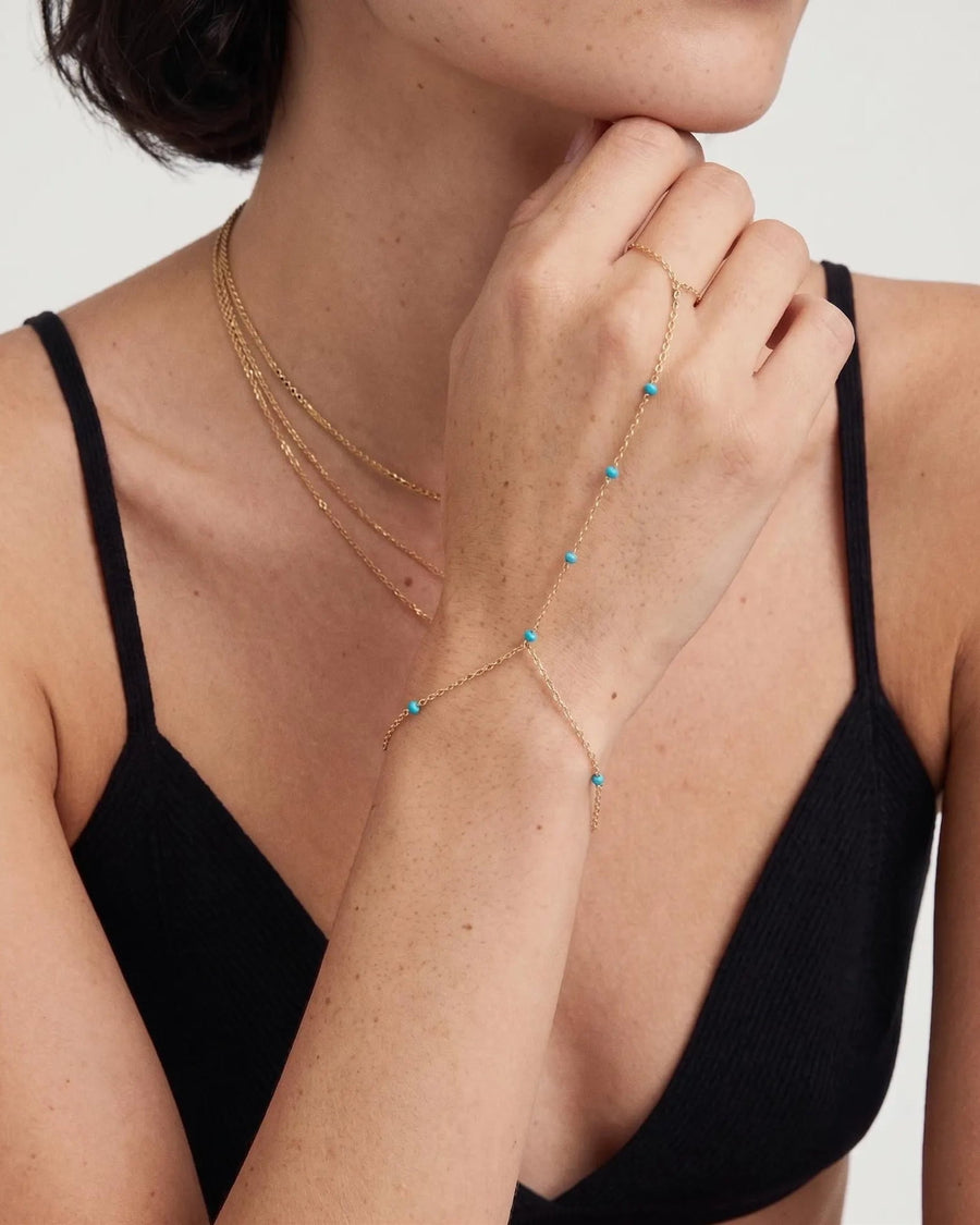 model image - A close-up of a model wearing a delicate gold chain bracelet with small turquoise blue bead accents, styled over a black ribbed camisole top against a light gray background.
